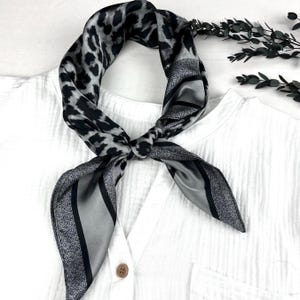 May include: A black and gray silk scarf with a leopard print pattern and striped design. The scarf is tied in a knot and is draped over a white shirt. The scarf is a stylish accessory.