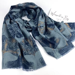 May include: A blue scarf featuring a dark blue and metallic gold ginkgo leaf pattern with frayed edges. A white card with the text Wrapped up By is partially visible in the upper right corner of the frame, resting near the fabric.