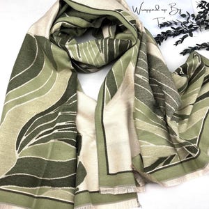 May include: A patterned scarf in shades of green and beige, with a fringed edge. The scarf features an abstract design and the text "Wrapped up By T" is visible.