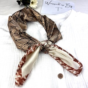Pode incluir: Um lenço de seda com um padrão de folhas em tons de castanho e preto, amarrado. O lenço tem uma borda creme com um desenho em zigue-zague castanho. O texto "Wrapped up By T" é visível no fundo.