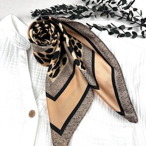 Può includere: Una sciarpa di seta arrotolata con un motivo leopardato. La sciarpa presenta uno sfondo beige con macchie di leopardo nere e un bordo nero. La sciarpa è drappeggiata su una camicia bianca.
