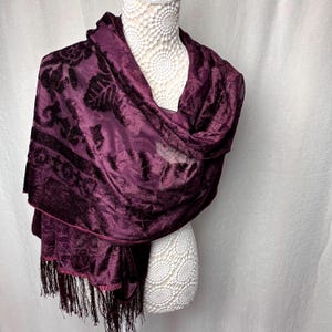 May include: A deep purple shawl with a floral pattern and fringe. The shawl is draped over a white mannequin. The fabric has a velvet texture, and the fringe is a darker shade of purple. The shawl is a stylish accessory.