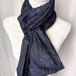 Op de afbeelding: Een marineblauwe sjaal met een bloemmotief en glinsterende accenten. De sjaal is over een witte mannequin gedrapeerd, wat de textuur en het ontwerp benadrukt. De stof oogt zacht en elegant, geschikt voor diverse gelegenheden.