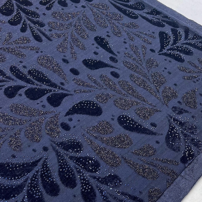 May include: A close-up shot of a navy blue fabric featuring a repeating pattern of stylized leaf shapes. The leaves are a darker navy with a velvet texture and are embellished with tiny, sparkling silver dots. The background is a solid navy blue.