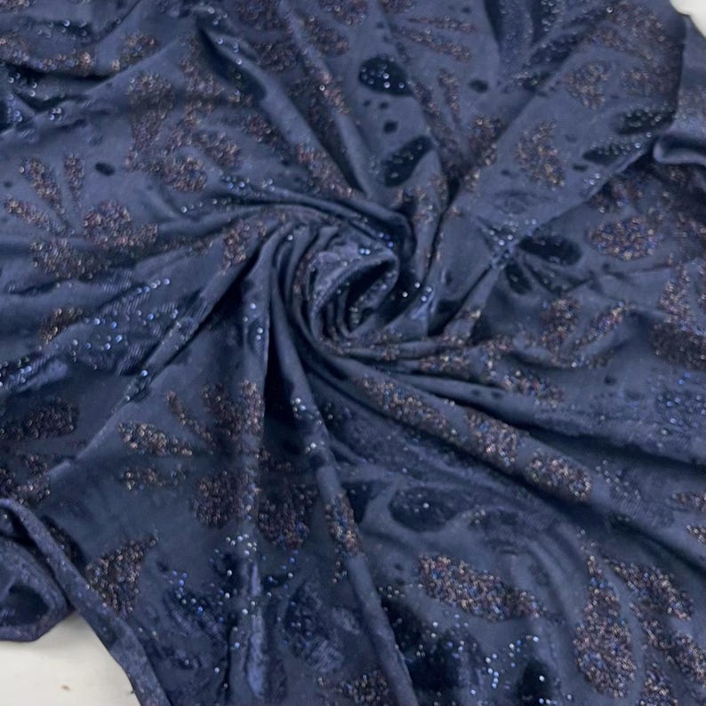 May include: Dark blue fabric with a velvet texture and a swirling pattern. The fabric features scattered sequins and a darker, textured design. This luxurious, shimmering textile is suitable for clothing or home decor projects.