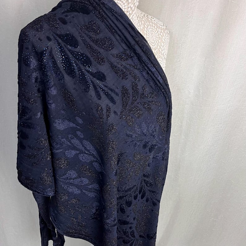 May include: A navy blue shawl with a floral pattern. The fabric has a velvet texture with shimmering accents. The shawl is draped over a mannequin, showcasing its design and drape. The floral design is in a darker shade of blue.