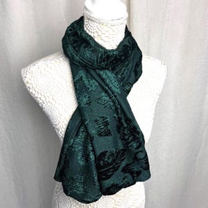 Op de afbeelding: Een donkergroene sjaal met een bladerenpatroon en een subtiele glans is over een witte mannequin gedrapeerd. De stof van de sjaal heeft een textuur, met donkerdere groene bladvormen tegen een lichtere groene achtergrond. De sjaal is een stijlvol accessoire.