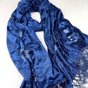 Op de afbeelding: Een vloeiende, marineblauwe sjaal met een fluwelen textuur en een bloemenpatroon. De sjaal heeft een doorschijnende, bedrukte overlay en is afgewerkt met franjes langs de randen. De sjaal is gedrapeerd en afgebeeld tegen een witte achtergrond.