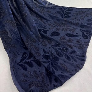 May include: Navy blue fabric featuring a repeating pattern of dark, glittery leaf shapes. The fabric is draped, with folds and shadows that highlight the texture and design. The overall effect is elegant and luxurious.