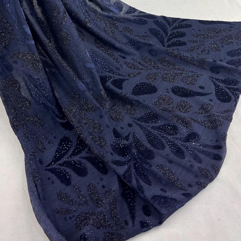 May include: Navy blue fabric featuring a repeating pattern of dark, glittery leaf shapes. The fabric is draped, with folds and shadows that highlight the texture and design. The overall effect is elegant and luxurious.