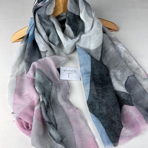 May include: A patterned scarf in a watercolour style, featuring grey, blue, and pink hues. The scarf is displayed on a wooden hanger. A small white tag with the text "Wrapped Up By T" is visible.