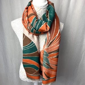 May include: A patterned scarf in shades of orange, green, brown, and beige. The scarf is decorated with a floral design. It is displayed on a white mannequin. The scarf has a fringed edge. The scarf's design is reminiscent of a flower.