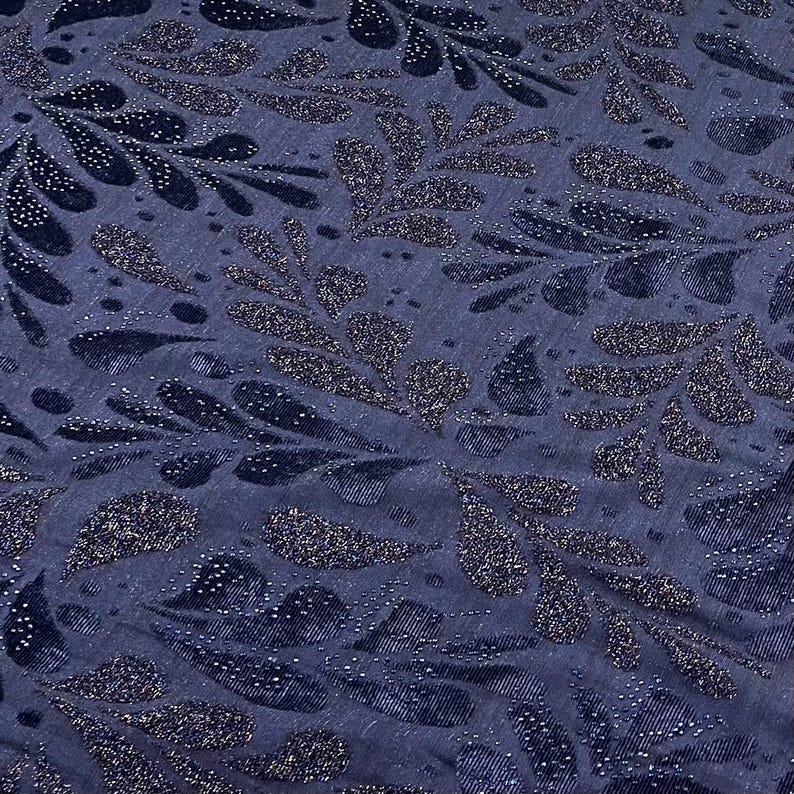 May include: Close-up of a navy blue fabric featuring a repeating pattern of stylised leaves. The leaves are embellished with glitter, creating a shimmering effect. The fabric has a textured appearance, with subtle vertical lines.
