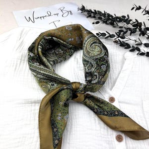 Può includere: Una sciarpa di seta con un motivo paisley nei toni del verde oliva, marrone e bianco è annodata. La sciarpa è drappeggiata su una camicia bianca. Il testo "Wrapped up By T" è visibile su una carta bianca.