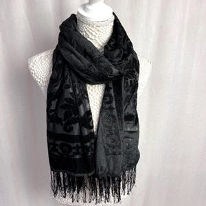 May include: Black velvet scarf with a floral pattern and fringe. The scarf is draped over a white mannequin bust. The fabric has a soft, textured appearance. The scarf's design includes a repeating floral motif.