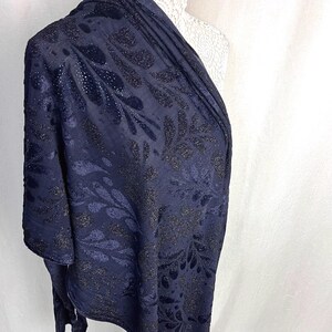 May include: Navy blue shawl with a floral pattern. The fabric has a textured appearance with shimmering accents. The design features dark, stylized leaves and floral shapes, creating a sophisticated look. The shawl is draped over a mannequin.