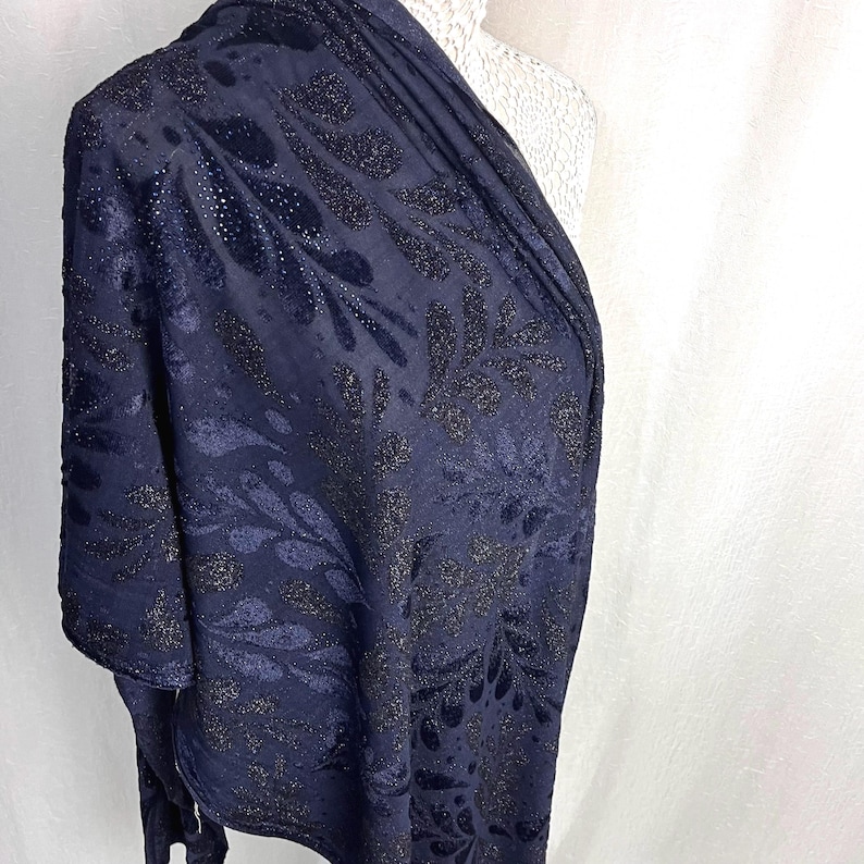 May include: Navy blue shawl with a floral pattern. The fabric has a textured appearance with shimmering accents. The design features dark, stylized leaves and floral shapes, creating a sophisticated look. The shawl is draped over a mannequin.