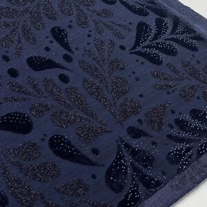 May include: A close-up shot of a navy blue fabric with a repeating pattern of stylized leaves and teardrop shapes. The design features a velvet texture with sparkling glitter accents, creating a luxurious appearance. The fabric has a subtle sheen.