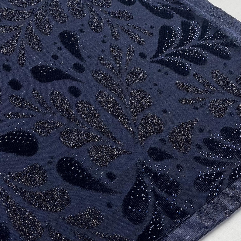 May include: A close-up shot of a navy blue fabric with a repeating pattern of stylized leaves and teardrop shapes. The design features a velvet texture with sparkling glitter accents, creating a luxurious appearance. The fabric has a subtle sheen.