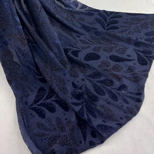 May include: Navy blue fabric with a repeating pattern of dark, glittery leaf shapes. The fabric is draped, showing folds and shadows, and has a soft, velvety texture. The design is visible against a lighter background.