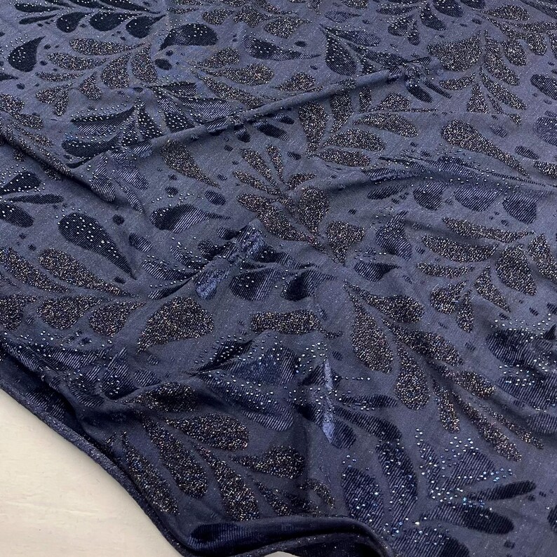 May include: Navy blue fabric with a shimmering, leaf-like pattern. The design features dark blue and silver glitter accents, creating a textured and elegant appearance. The fabric is folded, showing its drape and texture.