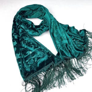 May include: A teal green velvet scarf with a floral pattern and fringe trim. The scarf is draped and folded, showcasing the soft texture and rich colour. The fringe hangs along the bottom edge.