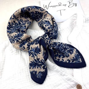 Puede incluir: Un pañuelo de seda con un estampado floral en azul marino y beige. El pañuelo está anudado y descansa sobre una camisa blanca. El texto "Wrapped up By T" es visible en el fondo.