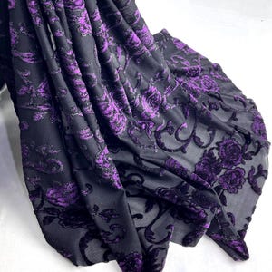 Op de afbeelding: Zwarte stof met een paars bloemenpatroon. De stof heeft een fluwelen textuur en een doorschijnend uiterlijk. Het bloemmotief omvat rozen en andere decoratieve elementen. De stof is gedrapeerd en toont zijn textuur en ontwerp.