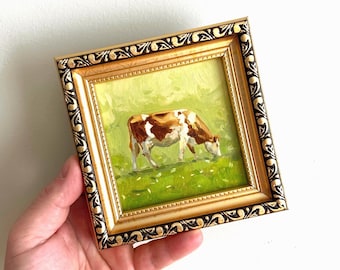 May include: A framed miniature painting of a cow grazing in a green field. The cow is brown and white. The frame is gold with a decorative black border. The painting is held by a hand.