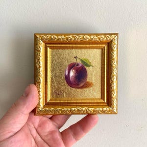 May include: A miniature painting in a gold frame, depicting a plum with a green leaf. The plum is a rich purple hue, set against a gold background. The frame is square and features an elaborate, decorative design.