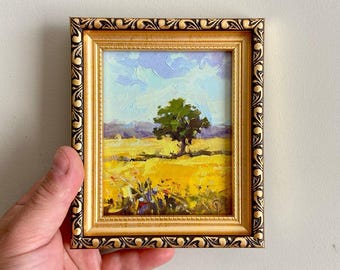 Op de afbeelding: Een klein olieverfschilderij in een decoratieve gouden lijst met een landschap met een enkele groene boom in een felgeel veld onder een lichtblauwe en paarse lucht. De lijst heeft een herhalend donkerbruin patroon langs de buitenrand.