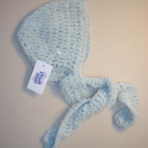 Op de afbeelding: Een lichtblauwe gehaakte baby muts met een bijpassende sjaal. De muts heeft een puntige top en de sjaal is in een knoop geknoopt.