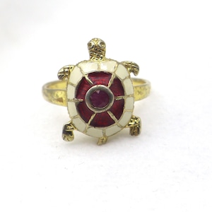 Puede incluir: Anillo de tortuga dorado con caparazón de esmalte rojo y blanco y una pequeña gema roja redonda en el centro. La cabeza y las patas de la tortuga están detalladas, y la banda del anillo es de oro pulido.