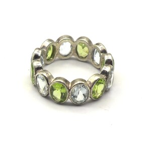 May include: A silver eternity band ring featuring a repeating pattern of alternating oval-cut light green and clear white gemstones set in a continuous metal frame. The ring has a polished metallic finish and a uniform circular shape.