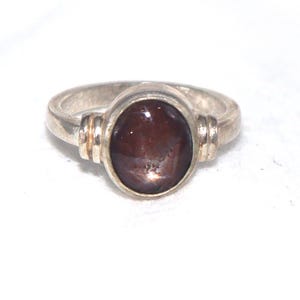 May include: A silver ring featuring a smooth, oval-shaped dark reddish-brown cabochon gemstone set in a bezel. The band has a polished metallic finish with two horizontal ridges on each side of the central stone setting, creating a simple design.