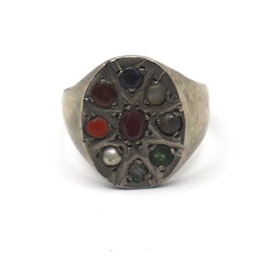 May include: A silver-toned metal ring featuring an oval face with a star-shaped cutout pattern. The ring is set with eight small, round, multi-colored cabochon stones arranged in a circle around a single central dark red stone.