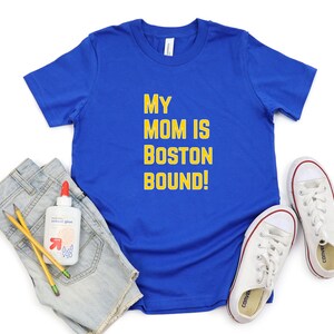 May include: A royal blue t-shirt with the text "MY MOM IS BOSTON BOUND!" in yellow. The shirt is paired with denim shorts, white trainers with red accents, pencils, and school glue.