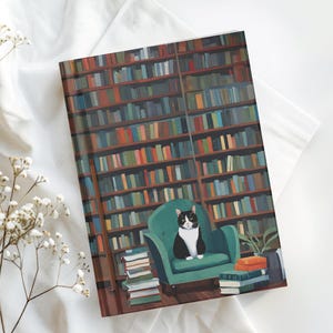 May include: A notebook with a library scene illustration. The image features a black and white cat sitting in a teal armchair in front of a bookshelf filled with books. A stack of books and a small plant are also visible.