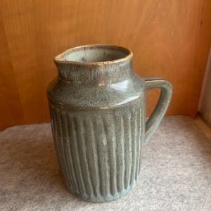Könnte beinhalten: Ein grüner Keramik-Krug mit einer strukturierten, gerippten Oberfläche und einem Henkel. Der Krug hat eine hellblaue Glasur mit einer gesprenkelten Oberfläche.