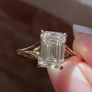 Może przedstawiać: Złoty pierścionek zaręczynowy z dużym, czystym diamentem o szlifie szmaragdowym.