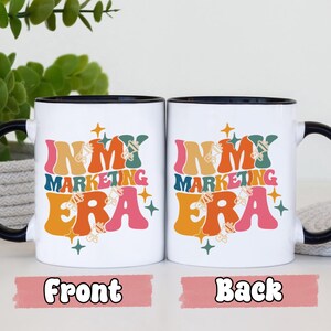 May include: White ceramic mug with a black rim and handle. The mug has a colorful graphic that says "In My Marketing Era" with a retro design. The front and back of the mug are shown.