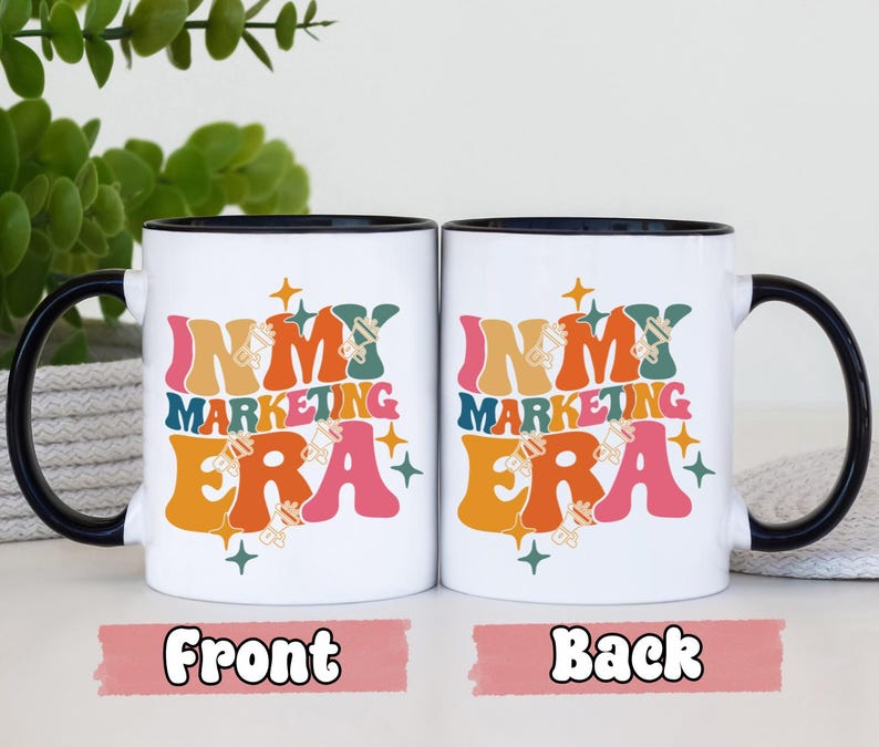 May include: White ceramic mug with a black rim and handle. The mug has a colorful graphic that says "In My Marketing Era" with a retro design. The front and back of the mug are shown.
