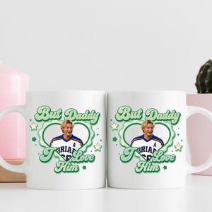 Puede incluir: Dos tazas de cer&aacute;mica blanca con el texto "But Daddy I Love Him" en verde y una foto de un hombre con una camiseta deportiva. Las tazas est&aacute;n decoradas con estrellas verdes y un borde en forma de coraz&oacute;n. Una vela rosa y un cactus est&aacute;n en el fondo.