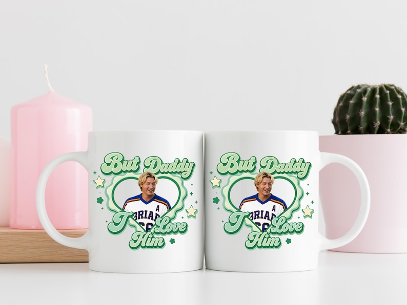 Puede incluir: Dos tazas de cer&aacute;mica blanca con el texto "But Daddy I Love Him" en verde y una foto de un hombre con una camiseta deportiva. Las tazas est&aacute;n decoradas con estrellas verdes y un borde en forma de coraz&oacute;n. Una vela rosa y un cactus est&aacute;n en el fondo.