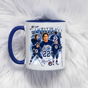 Puede incluir: Taza de cerámica blanca con asa e interior azules. La taza presenta un collage de imágenes de un jugador de hockey con uniforme azul con el nombre "Logan" y el número "22". También se ven las palabras "Meridian University Hawks".
