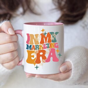 May include: White ceramic mug with a pink rim. The mug has a graphic design that says "In My Marketing Era" in colorful, retro-style lettering with stars and other design elements.