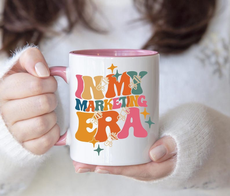 May include: White ceramic mug with a pink rim. The mug has a graphic design that says "In My Marketing Era" in colorful, retro-style lettering with stars and other design elements.