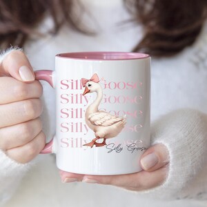 May include: White ceramic mug with a pink interior and handle. The mug features a watercolor illustration of a goose wearing a pink bow, with the words "Silly Goose" repeated in a pink font. A whimsical design.