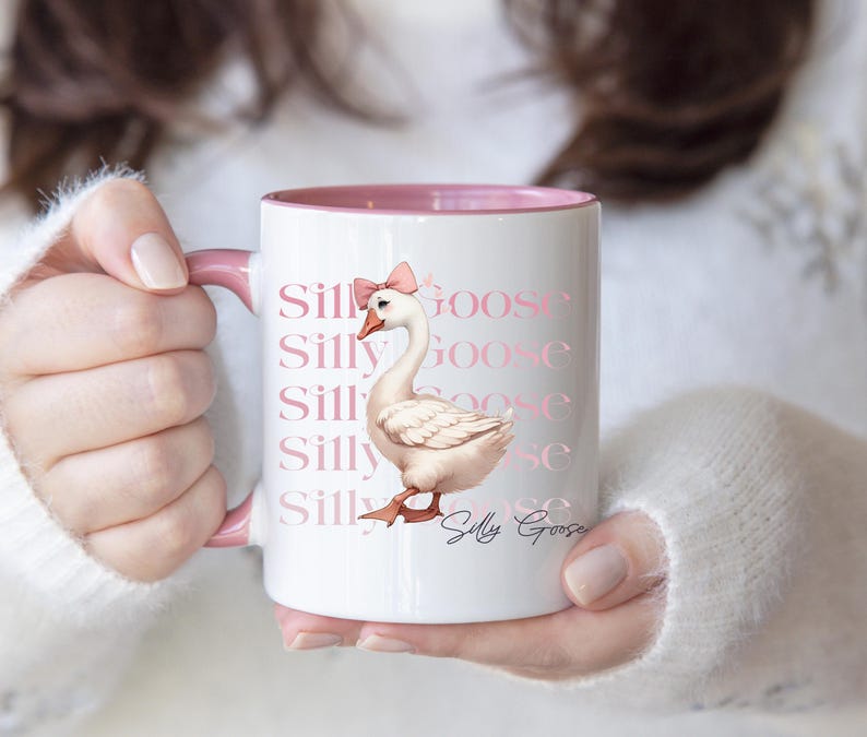 May include: White ceramic mug with a pink interior and handle. The mug features a watercolor illustration of a goose wearing a pink bow, with the words "Silly Goose" repeated in a pink font. A whimsical design.