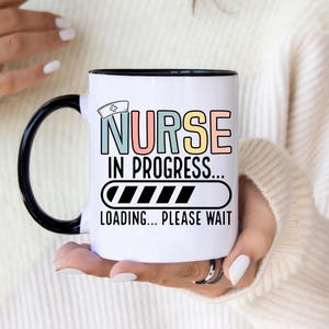 May include: White ceramic mug with a black handle and rim. The mug features the text "NURSE IN PROGRESS... LOADING... PLEASE WAIT" in pastel colours and a loading bar graphic, with a nurse's hat icon.